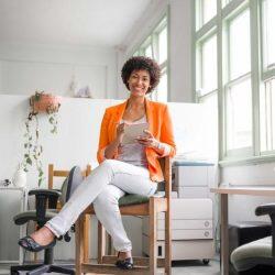 Woman With Orange Blazer On Sitting In An Office Setting Smiling